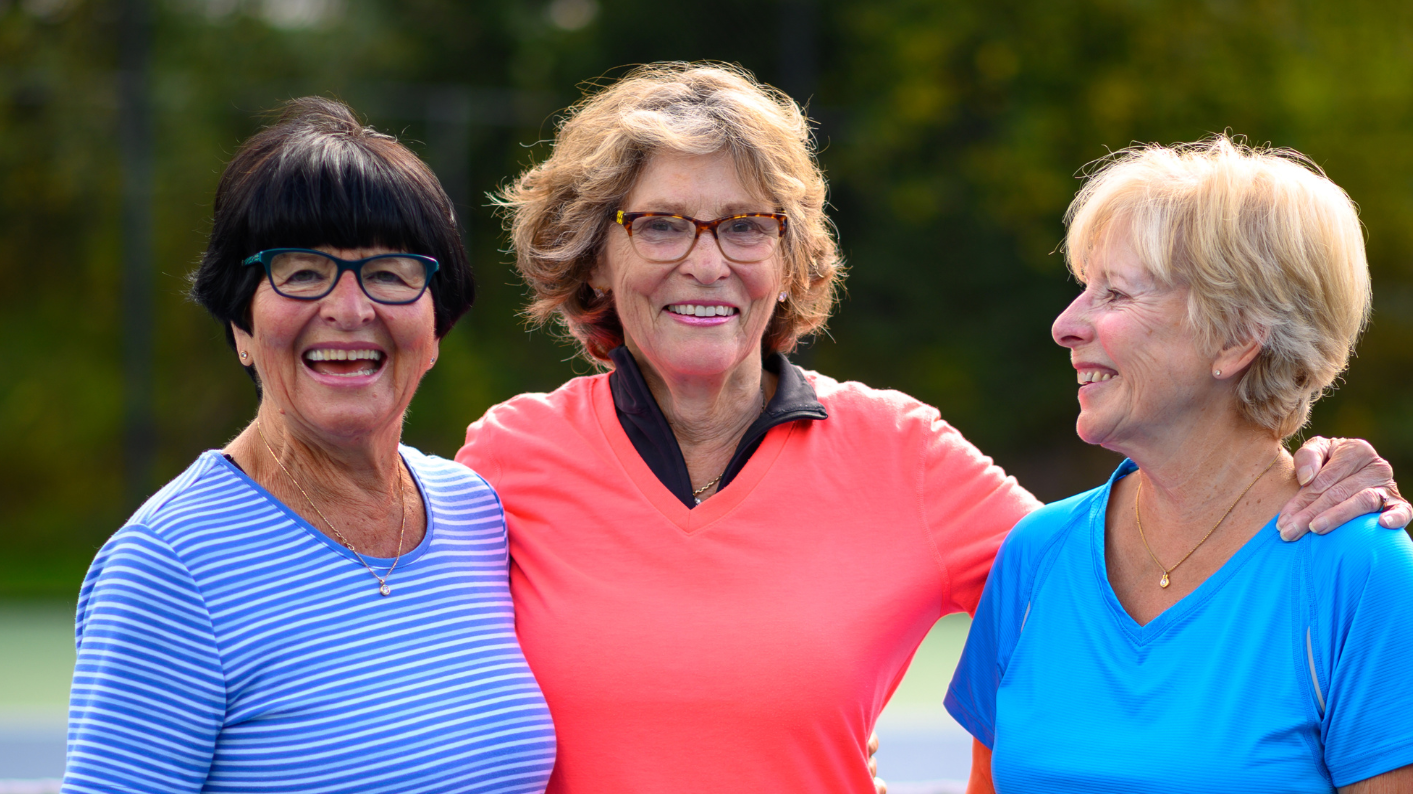 Tre glada kvinnliga seniorer står på en tennisplan och håller om varandra.