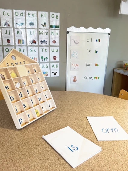 Bild på en tavla med bokstäver. I bakgrunden syns alfabetet och orden sil, is, ko och apa.