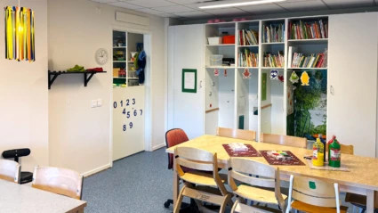 Inomhusmiljö med bord, stolar, böcker och pysselmaterial.