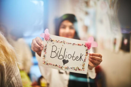 Ung tjej som håller i en skylt med texten "Bibliotek", pyntad med hjärtan och pärlor. Bilden är suddig av kort skärpedjup.