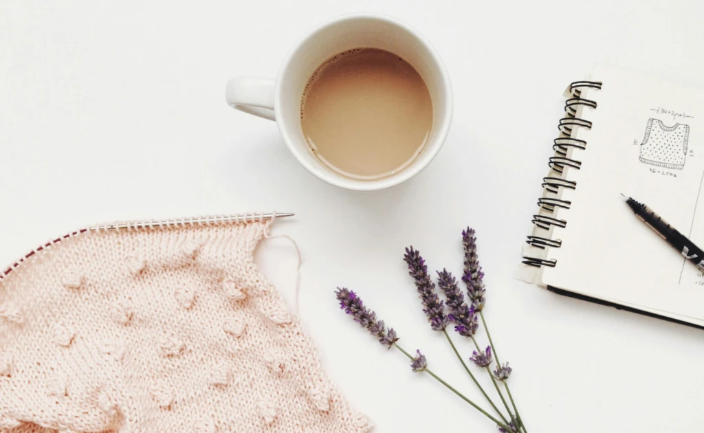 Ett vitt bord med en stickning med rosa garn, kaffekopp, lavendel och ett block med skiss på en väst.