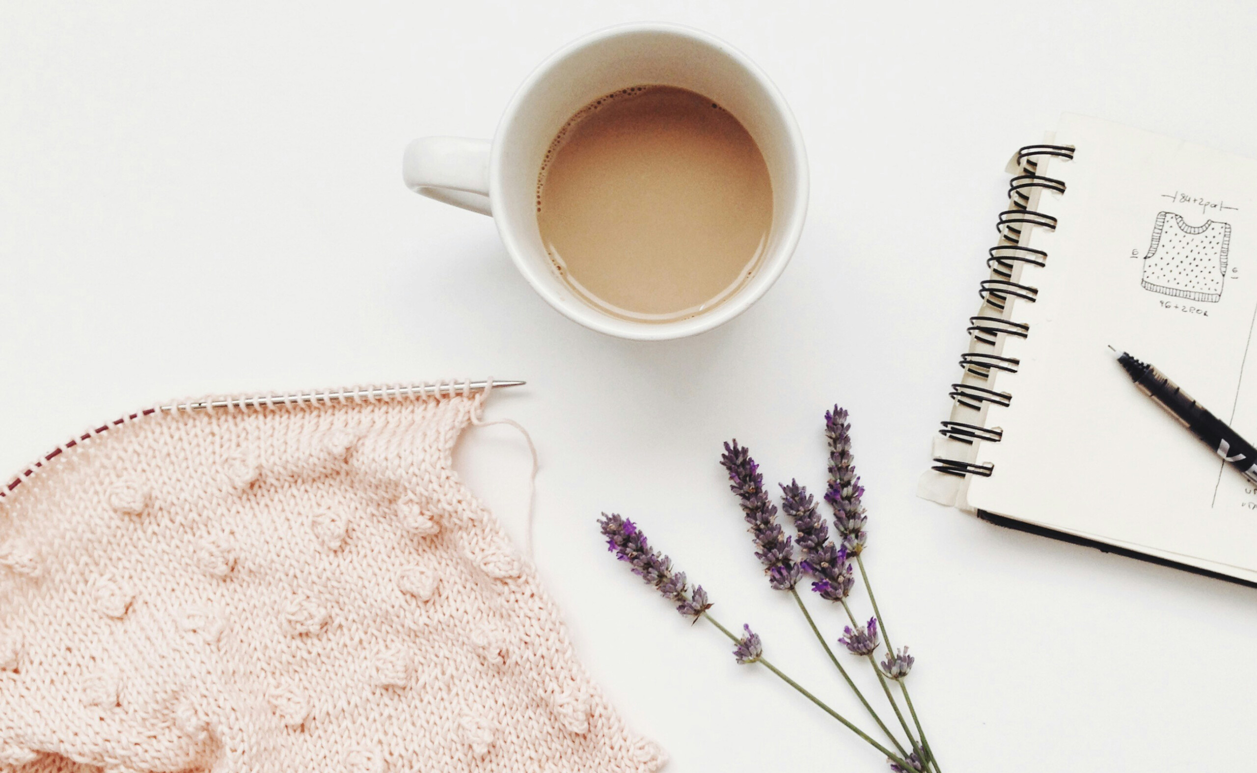 Ett vitt bord med en stickning med rosa garn, kaffekopp, lavendel och ett block med skiss på en väst.