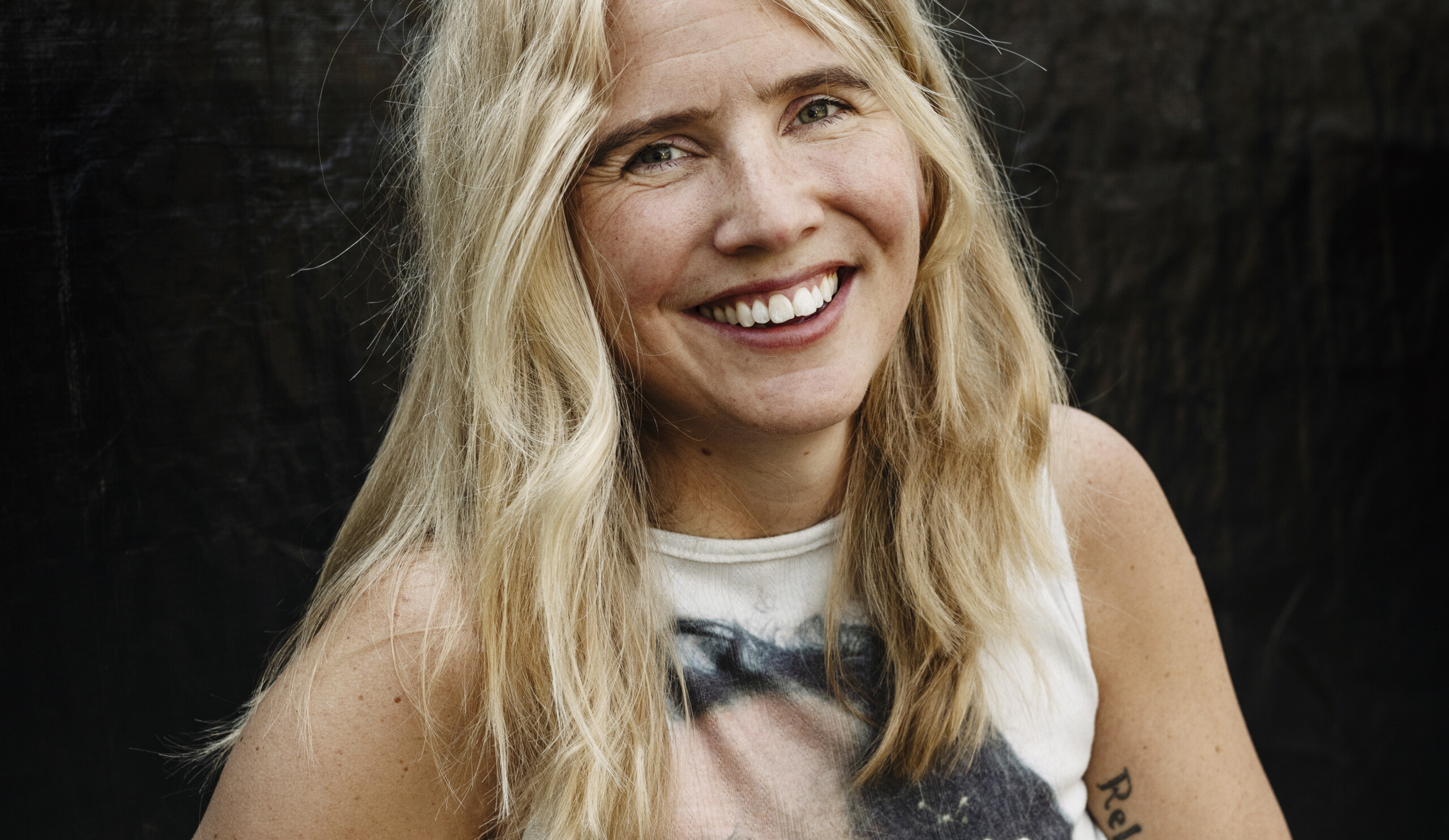 Porträtt av en leende Amanda Romare, med ljust linne och ljust hår, mot mörk bakgrund.
