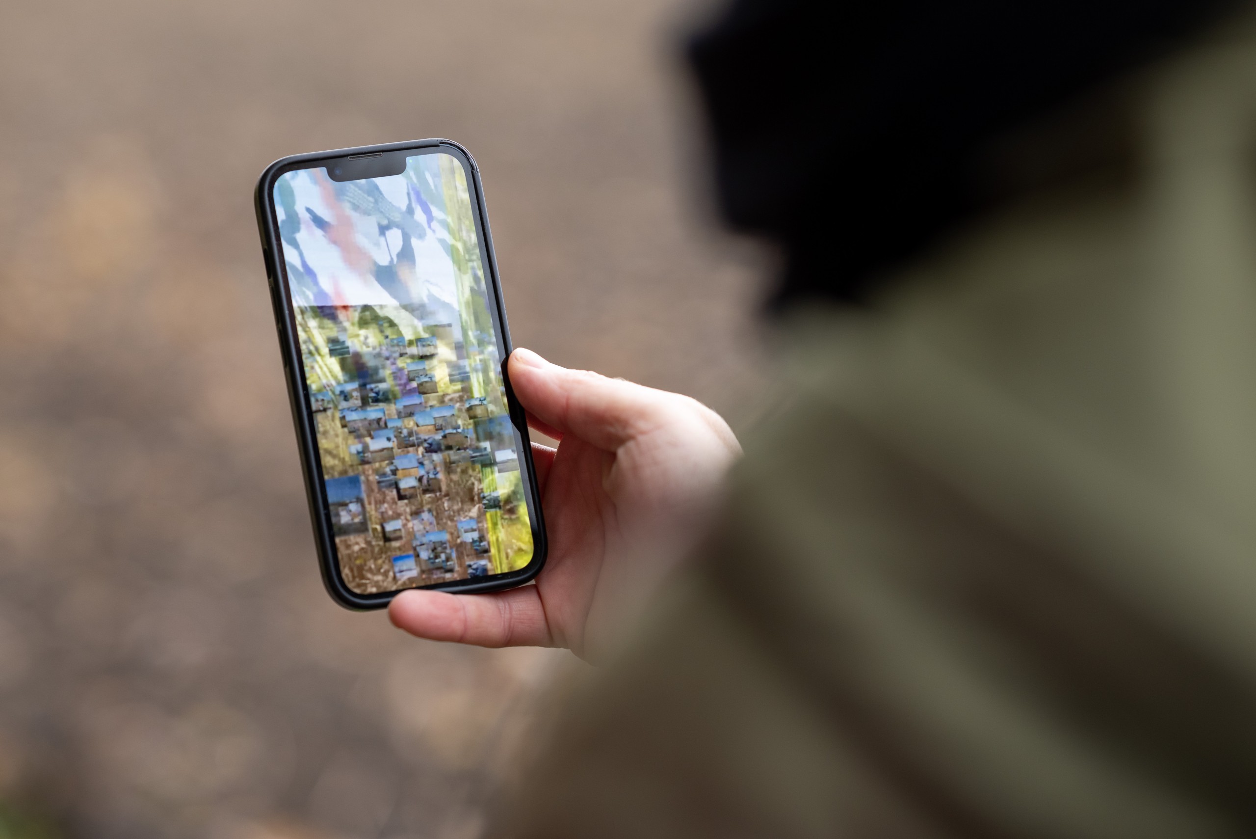 I fokus syns en mobiltelefon med pixlig skärm, som en person, ur fokus och sedd sett snett bakifrån håller i.
