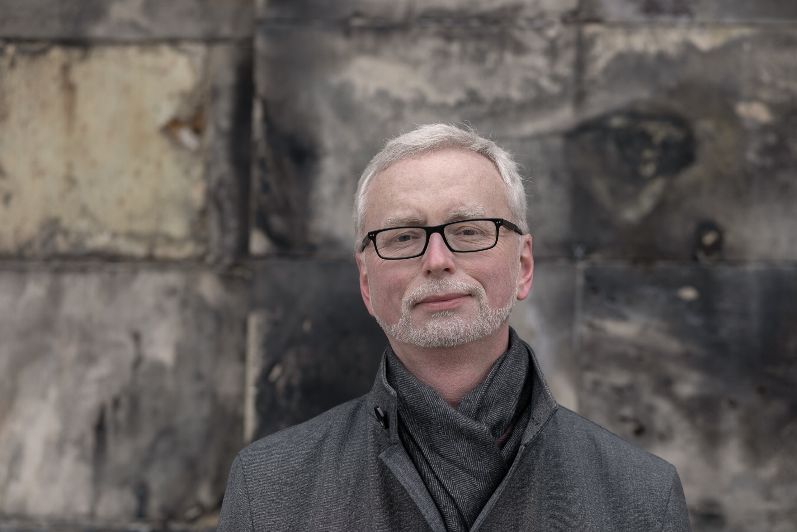 Porträtt av en man med svarta, stålbågade glasögon, grå rock och halsduk. Bakom skymtar några stenar av Lunds domkyrkas delvis svartnade kalkstensfasad.