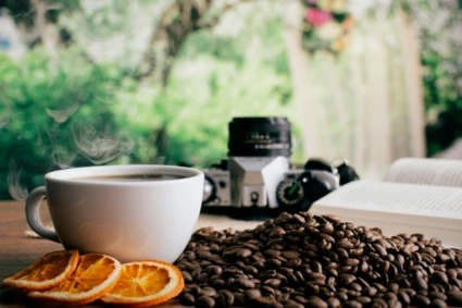 Kopp kaffe, kaffebönor, torkade apelsin skivor och en kamera mot en grön bakgrund.