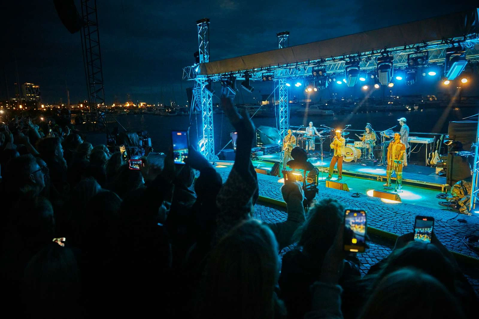 En konsert på en scen i havet med publik på kajen och blått ljus i natten.