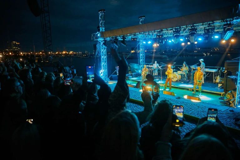 En konsert på en scen i havet med publik på kajen och blått ljus i natten.