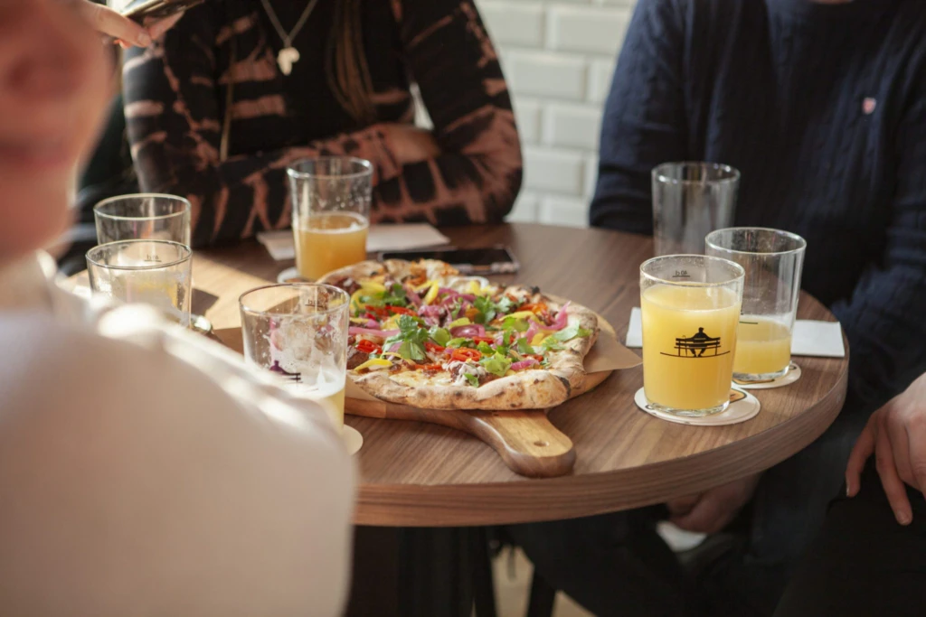 Närbild på ett bord där några personer sitter runt. I mitten står en nygräddad pizza och personerna har öl i deras glas.