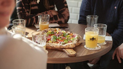 Närbild på ett bord där några personer sitter runt. I mitten står en nygräddad pizza och personerna har öl i deras glas.