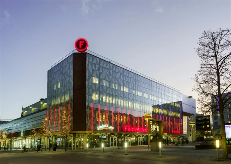 Kvällsbild över gallerian Söder. Hus med glaspartier formade som trianglar. Filmstadens F högst upp på taket.