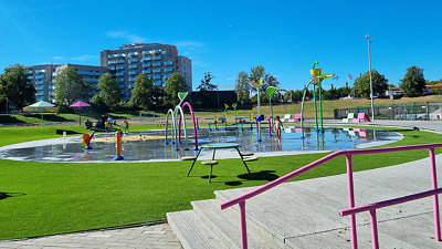 En vattenlekplats med olika delar som sprutar vatten på olika sätt. Runt lekplatsen en gräsmatta och trädäck.