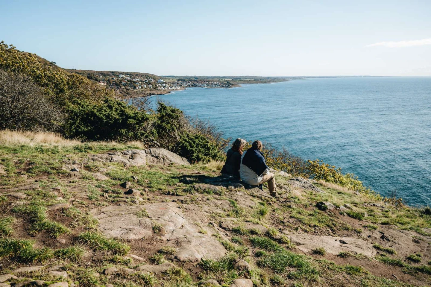 Ett par sitter uppe på Kullaberg och blickar ut över havet. Längre bort längs kusten skymtar Mölle.