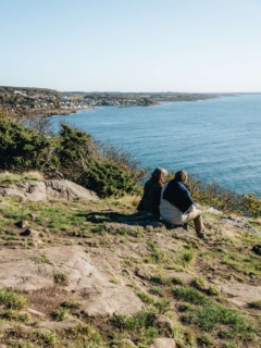 Ett par sitter uppe på Kullaberg och blickar ut över havet. Längre bort längs kusten skymtar Mölle.