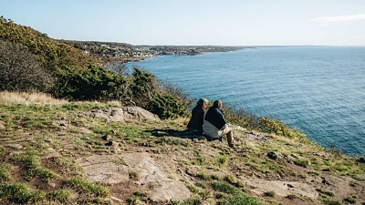 Ett par sitter uppe på Kullaberg och blickar ut över havet. Längre bort längs kusten skymtar Mölle.