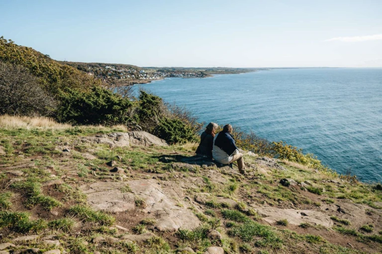 Ett par sitter uppe på Kullaberg och blickar ut över havet. Längre bort längs kusten skymtar Mölle.