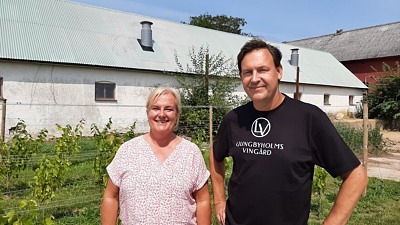 Bild på ägarparet till Ljungbyholms vingård, en man och en kvinna med gårdens längor och vinstockar i bakgrunden.