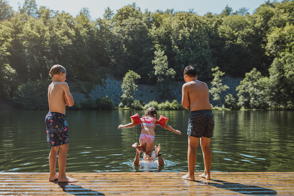 Bad i Odensjön i Söderåsens nationalpark. Sjön är omringad av grönskande natur. Två pojkar står på en brygga och ser när en mindre flicka hoppar ner i en vuxens famn i vattnet.