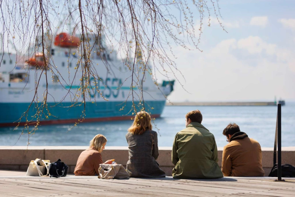 Ett gäng yngre personer sitter vid kajen och ser ut över hamnen där en forsea färja ligger. Vår i luften.