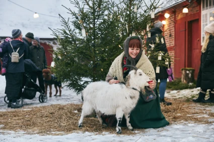 Kvinna i gammeldags klädsel sitter med en vit get och matar den med ett rött äpple