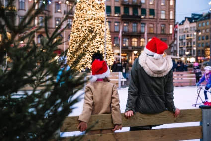 Två personer står och lutar sig med isrinken på isbanan. i bakgrunden syns en hög julgran med tusentals ljus tända.