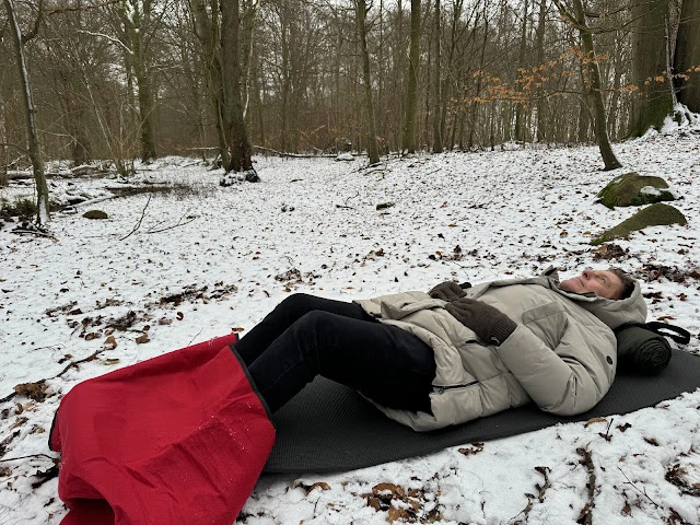 man ligger på ett liggunderlag på lätt snötäckt mark i skogen och tittar upp mot trädkronorna