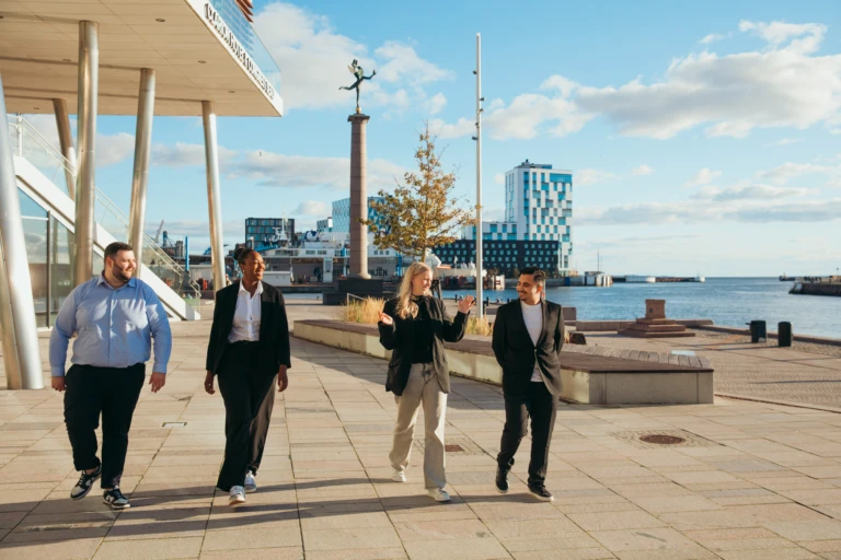 fyra personer samtalar och promenerar längs kajkanten med staden som siluett i bakgrunden.