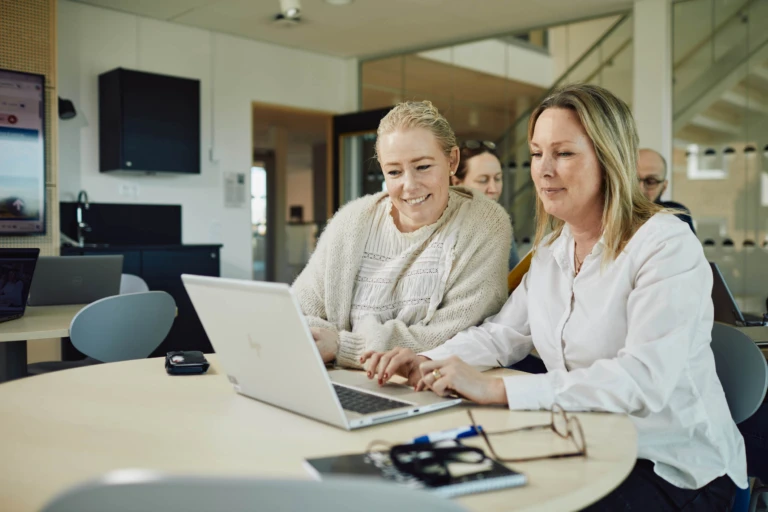 Två kvinnor samarbetar vid en laptop i ett klassrum.