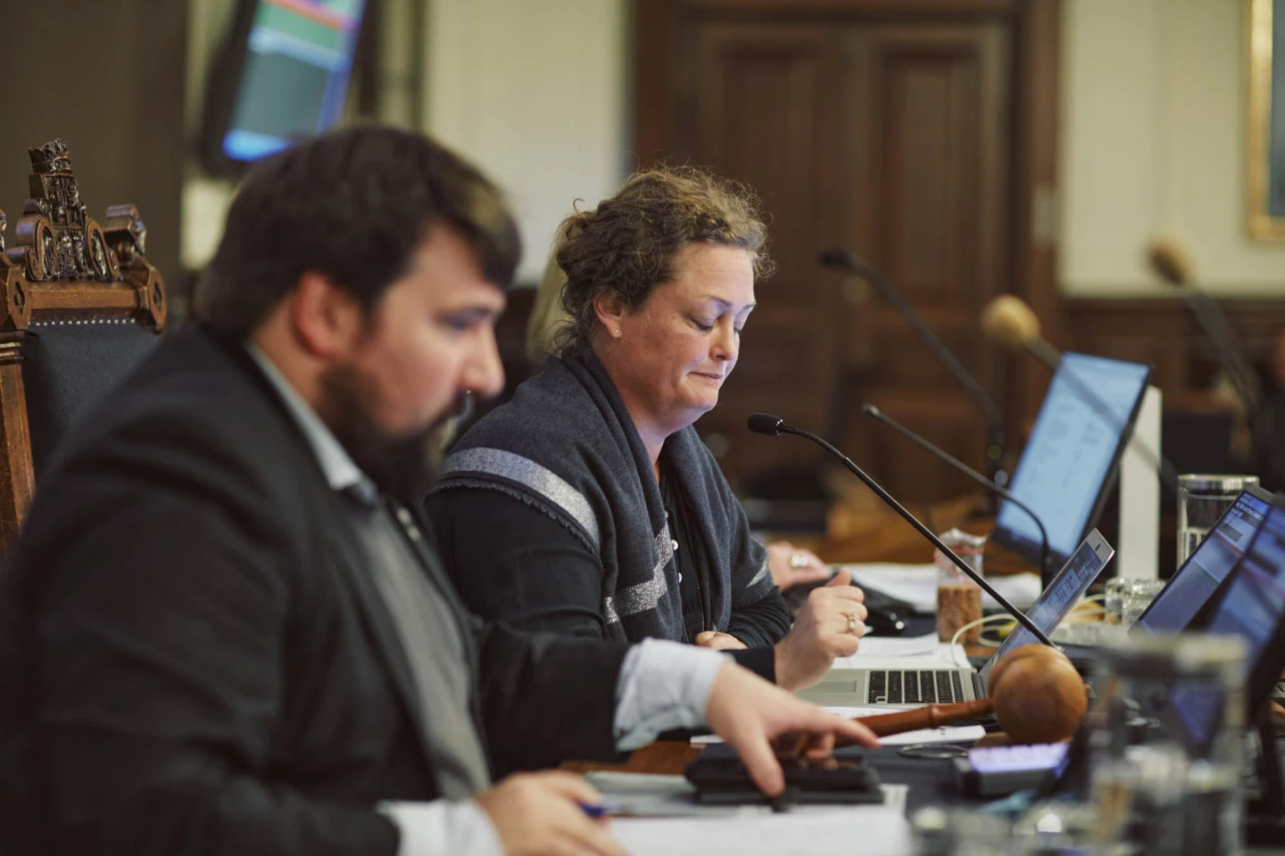 Två personer sitter vid ett bord med datorer och mikrofoner under ett formellt möte.