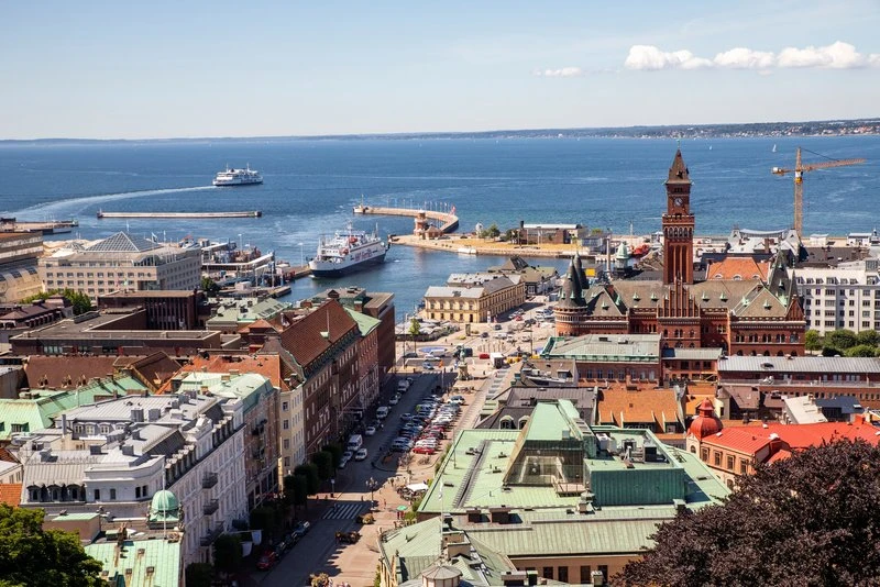 Utsikt över Helsingborgs hamn och stadskärna en solig dag, med färjor på väg ut mot havet och stadshuset med sitt röda torn i förgrunden.