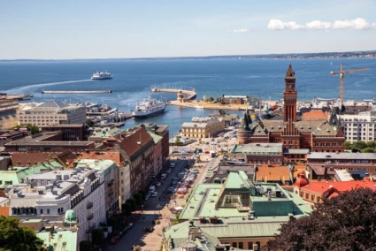 Utsikt över Helsingborgs hamn och stadskärna en solig dag, med färjor på väg ut mot havet och stadshuset med sitt röda torn i förgrunden.