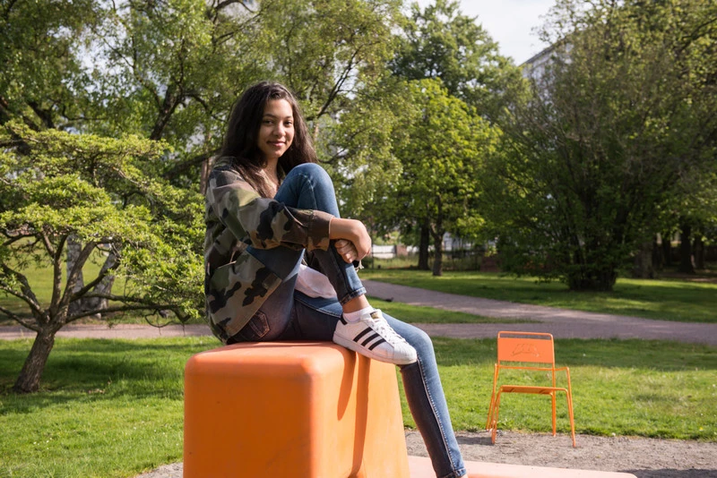 Ung kvinna sitter avslappnat på en orange betongkub i parken.