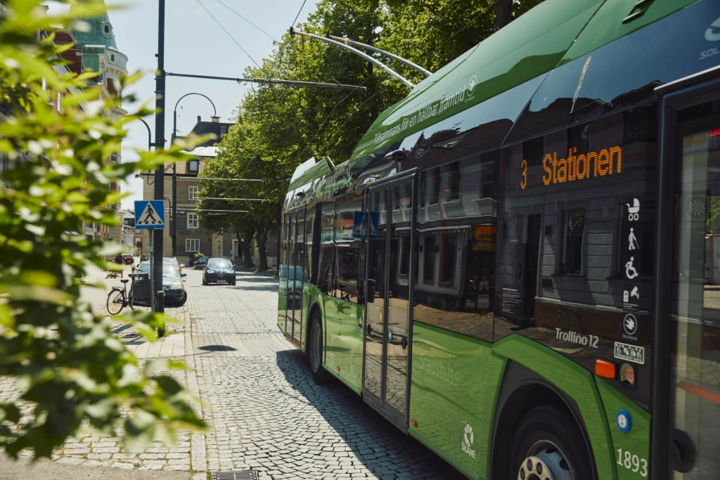 En grön stadsbuss kör på kullerstensgata i stadsmiljö.