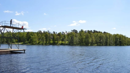 Solig sommardag vid Åsljungasjön där barn hoppar från hopptorn i det klara vattnet.