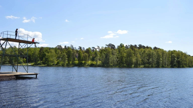 Solig sommardag vid Åsljungasjön där barn hoppar från hopptorn i det klara vattnet.
