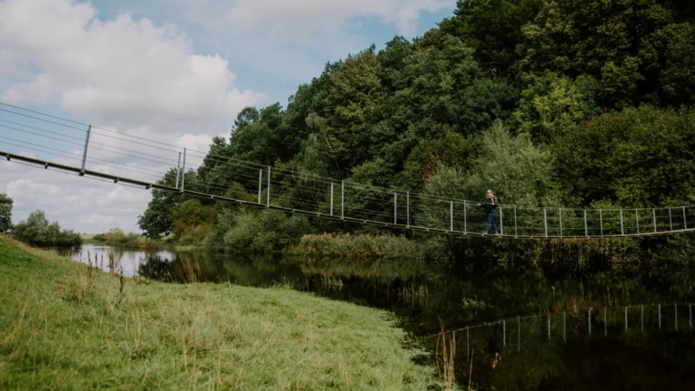 En smal hängbro som sträcker sig över ån vid Tomarp, inramad av lummig grönska.