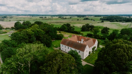 Bild av Tomarps kungsgård i Åstorps kommun, en historisk vit byggnad med rött tegeltak som ligger mitt i det skånska jordbrukslandskapet. Omgiven av träd, gräsmattor och öppna fält under en molnig sommardag, visar bilden både natur och kulturarv i harmoni.