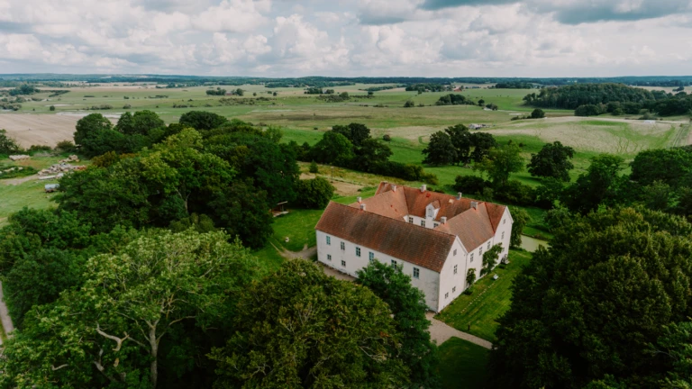 Bild av Tomarps kungsgård i Åstorps kommun, en historisk vit byggnad med rött tegeltak som ligger mitt i det skånska jordbrukslandskapet. Omgiven av träd, gräsmattor och öppna fält under en molnig sommardag, visar bilden både natur och kulturarv i harmoni.