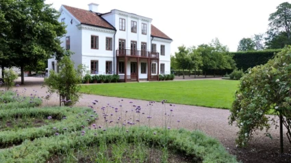 Bild av huvudbyggnaden på Fredriksdals museer och trädgårdar i Helsingborg. Den vita herrgårdsliknande byggnaden med rött tegeltak och träbalkong står mitt i en välskött trädgård med blomsterplanteringar, grusgångar och grönskande träd. Miljön utstrålar sommar och kulturhistoria i harmonisk parkmiljö.