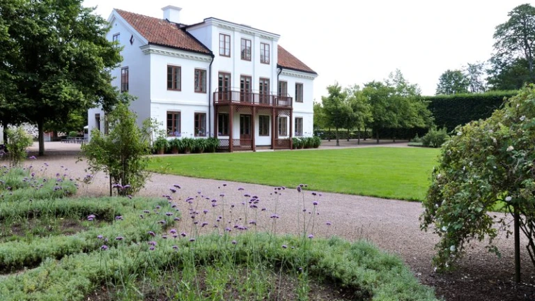 Bild av huvudbyggnaden på Fredriksdals museer och trädgårdar i Helsingborg. Den vita herrgårdsliknande byggnaden med rött tegeltak och träbalkong står mitt i en välskött trädgård med blomsterplanteringar, grusgångar och grönskande träd. Miljön utstrålar sommar och kulturhistoria i harmonisk parkmiljö.