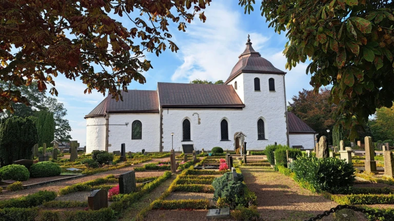 Bild av Gråmanstorps kyrka i Klippans kommun, en vitputsad byggnad med brunrött tak och torn i klassisk landsortsstil. Kyrkan ligger mitt i en prydlig kyrkogård med häckomgärdade gravplatser och gamla stenar. Gröna träd och höstfärgade löv ramar in motivet under en klar blå himmel.