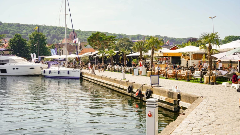 Sommardag i Båstads hamn där besökare njuter av mat och sol vid kajen bland båtar och palmer.