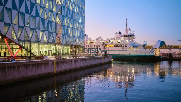 En ForSea-färja ligger förtöjd intill ett glasbeklätt kontorshus i Helsingborg medan ljusen speglar sig i vattnet.