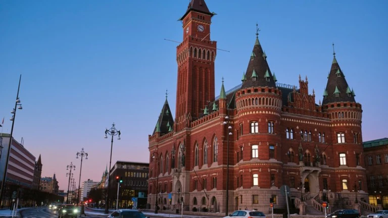 Bild av Helsingborgs rådhus, en pampig nygotisk byggnad i rött tegel med ett högt klocktorn och karakteristiska gröna koppartak. Fotot är taget i skymningen med bilar och gatlyktor längs Drottninggatan i förgrunden.