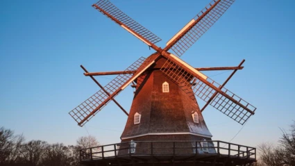 Bild av en klassisk väderkvarn i Helsingborg, upplyst av kvällssolen. Kvarnen är byggd i trä med flera små fönster och ett runt trädäck runt basen. Den står omgiven av träd under en klarblå himmel.