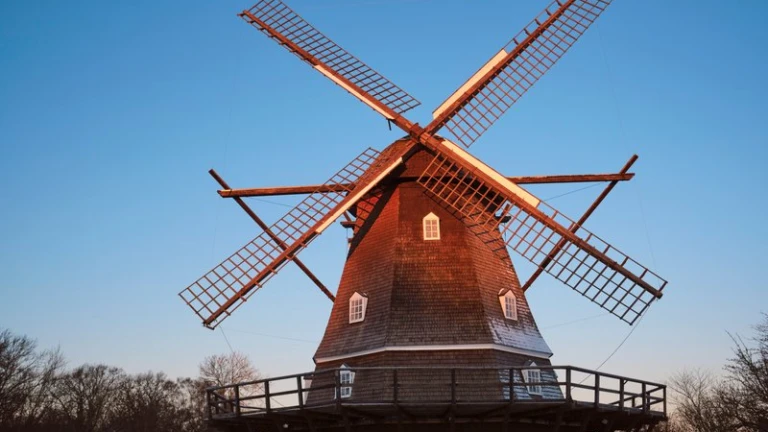 Bild av en klassisk väderkvarn i Helsingborg, upplyst av kvällssolen. Kvarnen är byggd i trä med flera små fönster och ett runt trädäck runt basen. Den står omgiven av träd under en klarblå himmel.