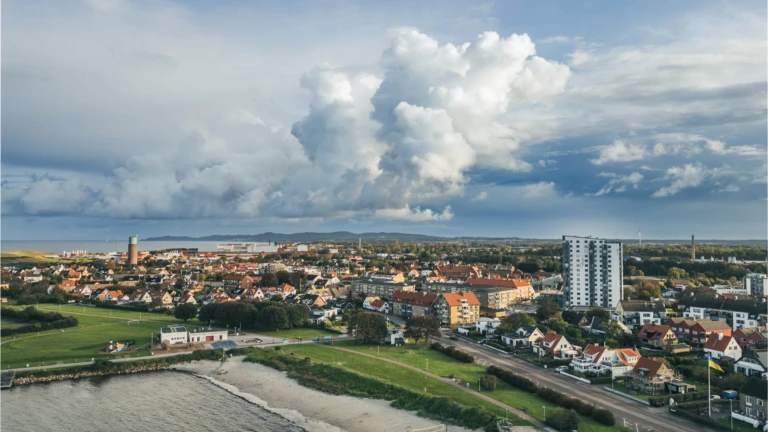 Utsikt över kuststaden Höganäs med strand, bostäder och hamn, inramad av dramatisk himmel och vida vyer.