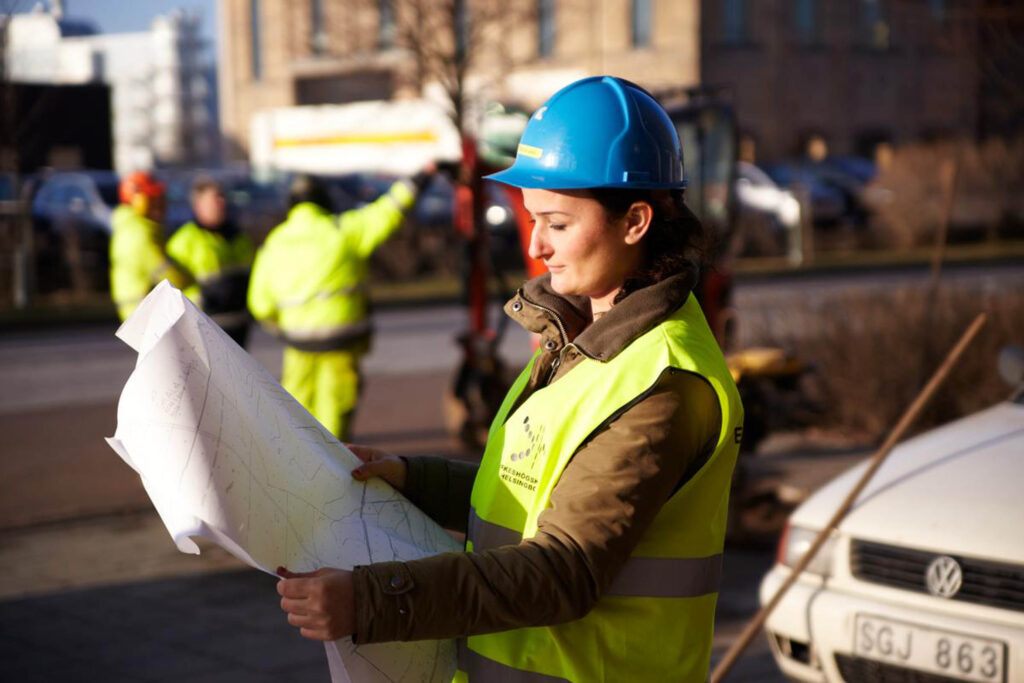 Kvinna i skyddshjälm och reflexväst som läser en ritning på en byggarbetsplats.