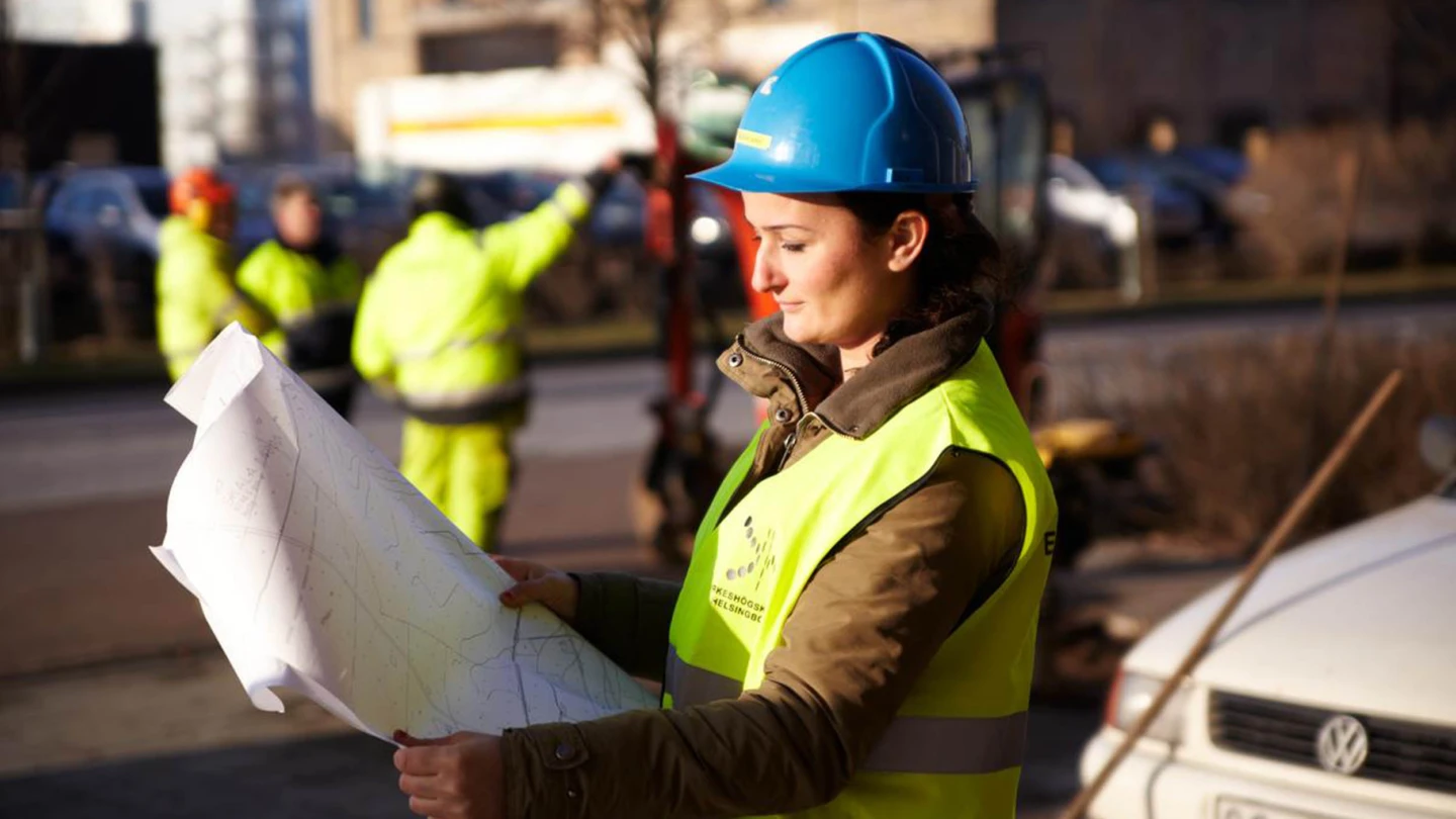 Kvinna i skyddshjälm och reflexväst som läser en ritning på en byggarbetsplats.
