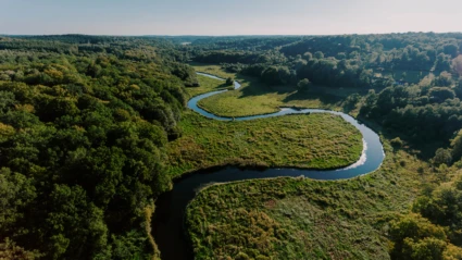 Från ovan syns en å som slingrar sig genom ett böljande landskap av skog och öppna fält i morgonljus.